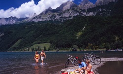 Eine Radlergruppe, die auf der Schweizer Seenroute unterwegs ist, macht an einem von Berggipfeln umgebenen See Pause. Ein Pärchen kommt gerade Hand in Hand aus dem Wasser, während ein anderes neben den abgestellten Rädern auf einem schmalen Kiesstrand sitzt.
