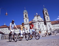 Drei junge Radlerinnen fahren auf der Schweizer Seenroute am markanten Kloster Einsiedeln vorbei.