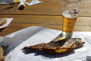 Ein auf einem undurchlässigen Papier gelegter Steckerlfisch und ein teilweise getrunkenes Bier auf einem Holztisch