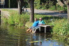 Zwei Radler machen an einem See Pause und lassen die Füße ins Wasser baumeln.