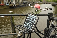 Ein abgestelltes Fahrrad auf einer Brücke über einem Kanal mit einer rosa Blume und einem Schild, das die Aufschrift "Happines ist not a destination, it is a way of life"
