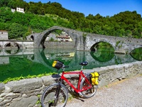 Ein rotes Fahrrad mit gelber Gepäck- und Lenkertasche steht bei Borgo Mazzano vor der Teufelsbrücke, die sich im grünen Wasser des Flusses Serchio spiegelt.