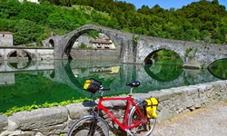 Ein rotes Fahrrad mit gelber Gepäck- und Lenkertasche steht bei Borgo Mazzano vor der Teufelsbrücke, die sich im grünen Wasser des Flusses Serchio spiegelt.