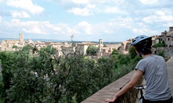 Ein Radfahrer steht in der Toskana an einer Steinmauer und schaut auf die vor ihm liegende Stadt.