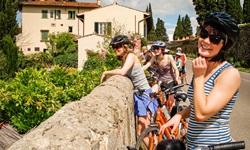 Mehrere Radlerinnen machen in einem toskanischen Dorf Pause und genießen, an eine Steinmauer gelehnt, die Aussicht.