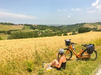 Radfahrerin sitzt neben ihrem orangenem Fahrrad im Gras, macht eine Pause und blickt über die schöne Landschaft der Toskana