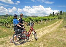 Zwei Radfahrer stehen neben ihren abgestellten Rädern auf einem Feldweg an einem Weinberg und blick über die Weinreben zur "Stadt der Türme", San Gimignano