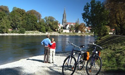 Ein Paar steht am Ufer der Donau und blickt zum Ulmer Münster, hinter sich sind ihre Räder abgestellt