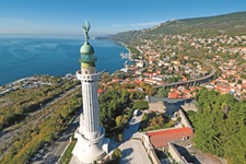 Luftaufnahme der von einem Engel gekrönten Siegessäule in Triest. Im Bildhintergrund sind die Dächer der Stadt und das tiefblaue Meer zu erkennen.