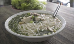 Ein Teller mit vietnamesischer Suppe, die unter anderem Glasnudeln und Fleisch enthält, steht auf einem hölzernen Tisch.