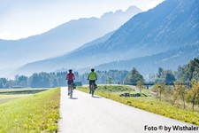 Zwei Radler fahren auf einem asphaltierten Abschnitt des Alpe-Adria-Radwegs auf eine wunderschöne Bergkulisse zu.