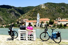 Ein Radfahrerpärchen macht auf einer Bank am Donauufer Pause und genießt den Ausblick auf Dürnstein.