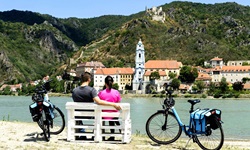 Ein Radfahrerpärchen macht auf einer Bank am Donauufer Pause und genießt den Ausblick auf Dürnstein.