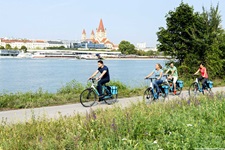 Eine Gruppe Radler auf dem Donauradweg bei Wien - am gegenüberliegenden Flussufer ist die Franz-von-Assisi-Kirche zu erkennen.
