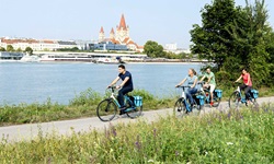 Eine Gruppe Radler auf dem Donauradweg bei Wien - am gegenüberliegenden Flussufer ist die Franz-von-Assisi-Kirche zu erkennen.
