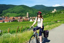 Eine Frau radelt auf einem Radweg entlang. Hinter ihr ist Kaysersberg mit seiner hoch thronenden Burg zu erkennen.