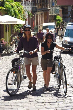 Zwei Frauen mit Helmen schieben ihre Räder durch ein Dorf im verlockenden Elsass