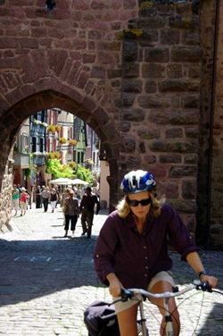 Blick auf ein ehemaliges Stadttor des verlockenden Elsass und deren dahinterliegende Fußgängerzone. Im Vordergrund radelt eine Frau.