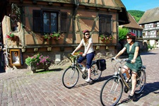 Blick auf zwei Fahrradfahrerinnen, die durch ein typisch elsässisches Dorf mit Kopfsteinpflaster und mit Blumen geschückten Fachwerkhäusern radeln