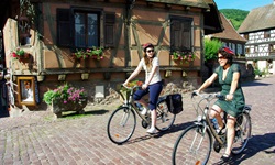Blick auf zwei Fahrradfahrerinnen, die durch ein typisch elsässisches Dorf mit Kopfsteinpflaster und mit Blumen geschückten Fachwerkhäusern radeln