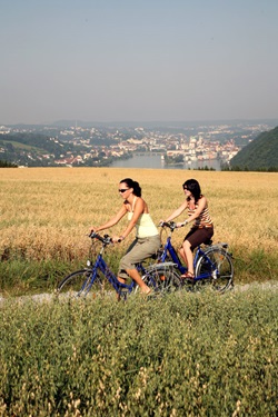 Zwei Frauen radeln auf dem Donauradweg, der sich hier zwischen zwei Getreidefeldern hindurchschlängelt. Im Hintergrund ist noch die Stadt Passau mit dem Drei-Flüsse-Eck zu erkennen.