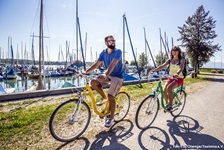 Ein Radfahrerpärchen passiert den Chiemsee, auf dem zahlreiche Boote schaukeln.