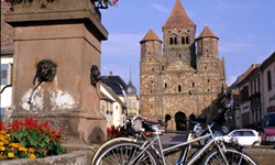 Zwei Fahrräder lehnen an einem mit Blumen geschmückten Brunnen im Elsass - dahinter ist ein Kloster zu erkennen