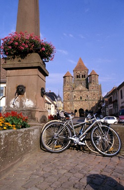 Zwei Fahrräder lehnen an einem mit Blumen geschmückten Brunnen im Elsass - dahinter ist ein Kloster zu erkennen