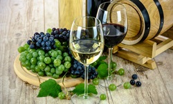 Zwei Weingläser - eines mit weißem, eines mit rotem Wein - vor einem Holzfässchen und neben einem Brett mit blauen und grünen Trauben.