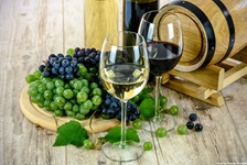Zwei Weingläser - eines mit weißem, eines mit rotem Wein - stehen vor einem Holzfässchen und neben einem Holzbrett mit blauen und grünen Trauben.