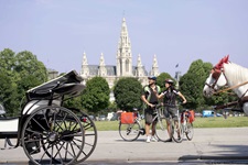 Zwei Radler sehen sich die wartenden Kutschen in Wien an, im Hintergrund sieht man das Wiener Rathaus