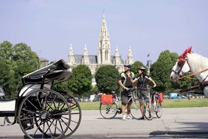 Zwei Radfahrer stehen auf einem Gehweg in Wien, während auf der Straße Kutschen stehen und im Hintergrund das Rathaus von Wien zu sehen ist