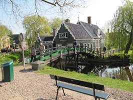 Eine Bank auf dem Weg an der Zaan mit Blick zu einer Brücke und den dahinterliegenden historischen Gebäude