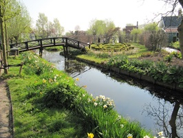 Eine Brücke über die Zaan mit Blick auf ein schön angelegtes Parkgelände und den dahinter liegenden typischen Holzhäusern der Zaanse Schans in Nordholland