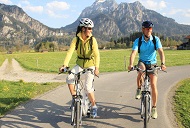 Zwei Radfahrer, die auf einer schmalen Landstraße durch eine grüne Landschaft mit Bergen im Hintergrund bei Füssen fahren.
