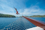 Eine Person springt vom Bug eines Bootes in das klare, blaue Meer, während in der Ferne sattgrüne Hügel zu sehen sind, Dalmatien, Kroatien