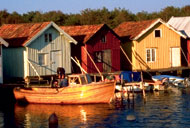 Reihe von bunten Fischerhütten aus Holz am Ufer von Saxemara, einem kleinen Fischerdorf in Südschweden. Im ruhigen Wasser liegt ein Holzboot vor Anker, das das traditionelle maritime Erbe der Region widerspiegelt.